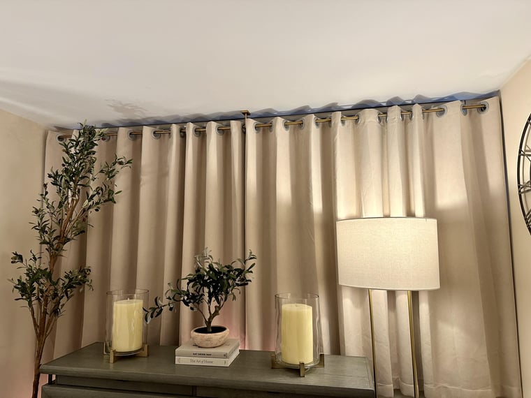 Beige blackout curtains with a brass rod, table with candles and plants, and a lamp in a room setting.