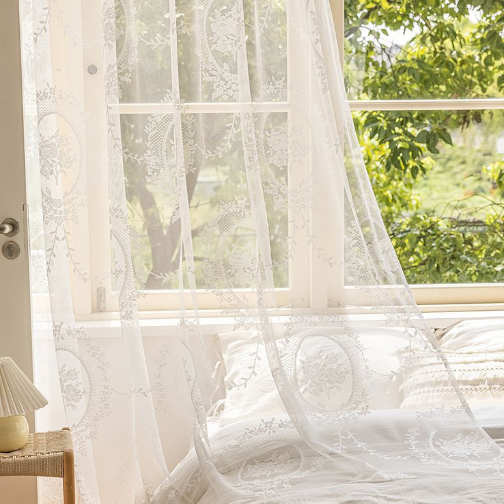 White sheer lace curtains with embroidered floral patterns hanging over a window in a bright bedroom, with white bedding and green foliage visible outside.