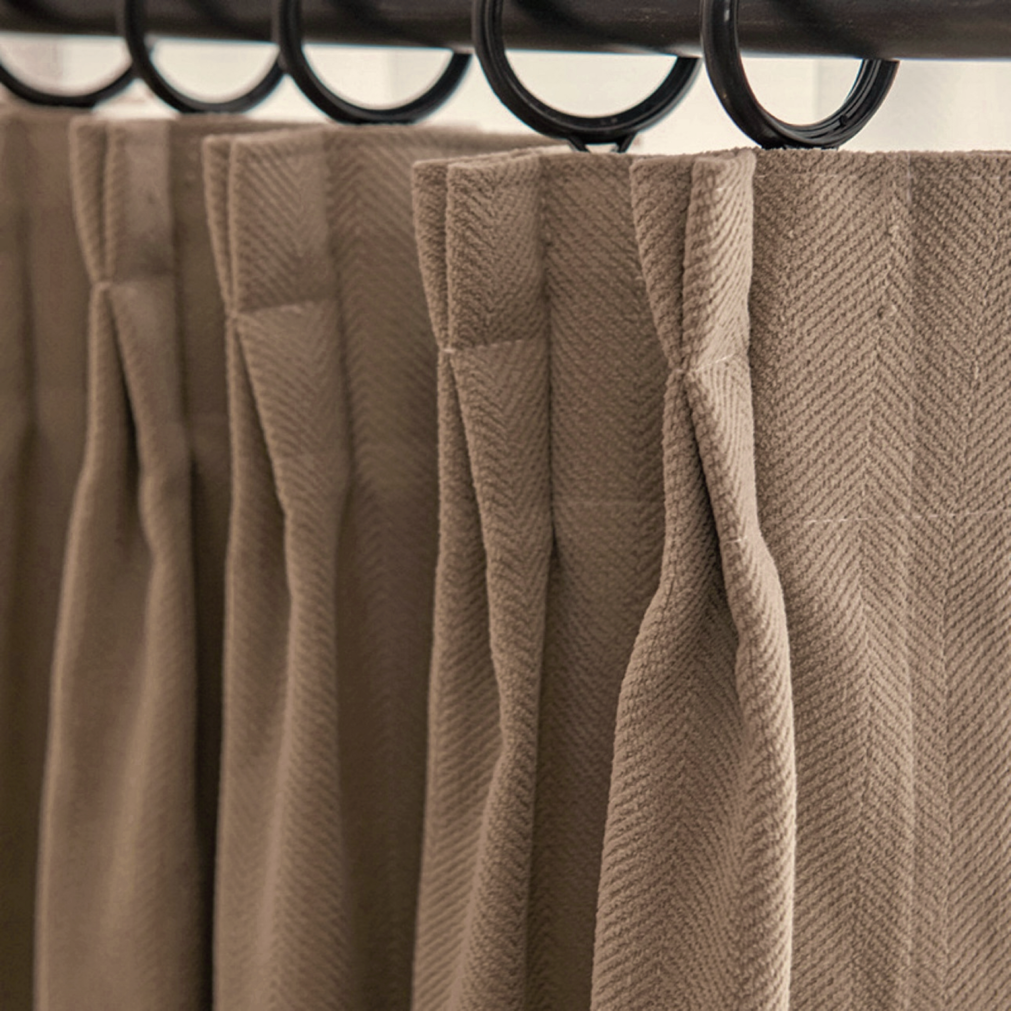 Close-up of brown textured fabric with pinch pleats and black rings on a rod.