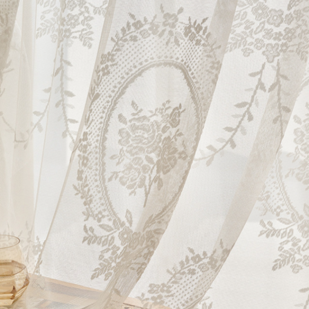 A close-up of a sheer white curtain with an elegant floral lace pattern in a soft ivory tone, draped elegantly with a glass and book visible in the softly lit background.
