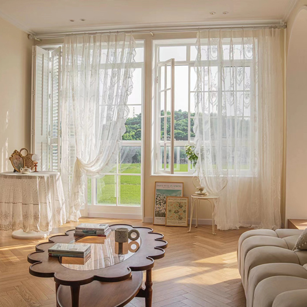 A bright, sunlit living room with tall sheer white curtains hanging over large windows, showing a green lawn outside. The room has warm cream walls, a scalloped wooden coffee table, and a light upholstered sofa.