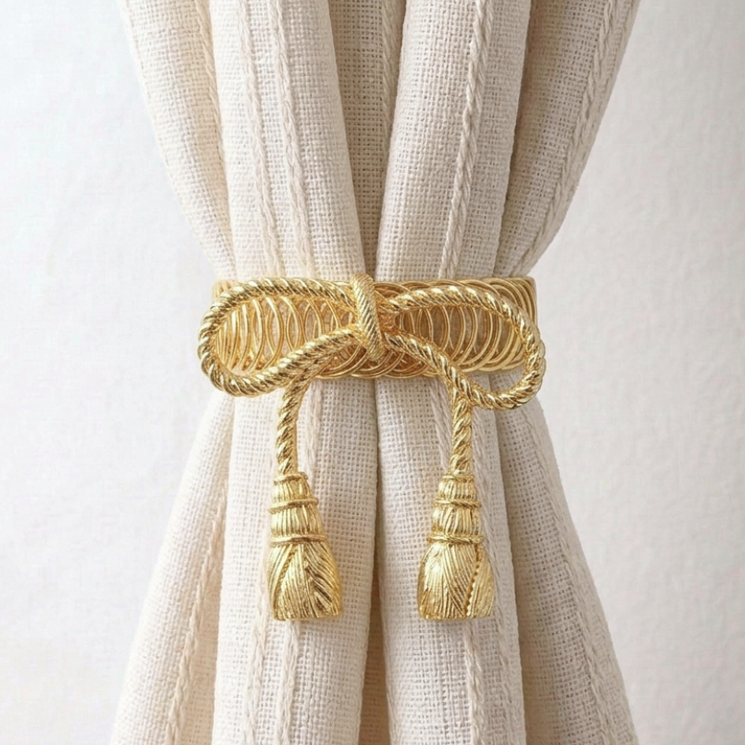 A close-up of a cream-colored linen curtain tied back with a decorative gold rope curtain tie and tassels, set against a plain off-white background.