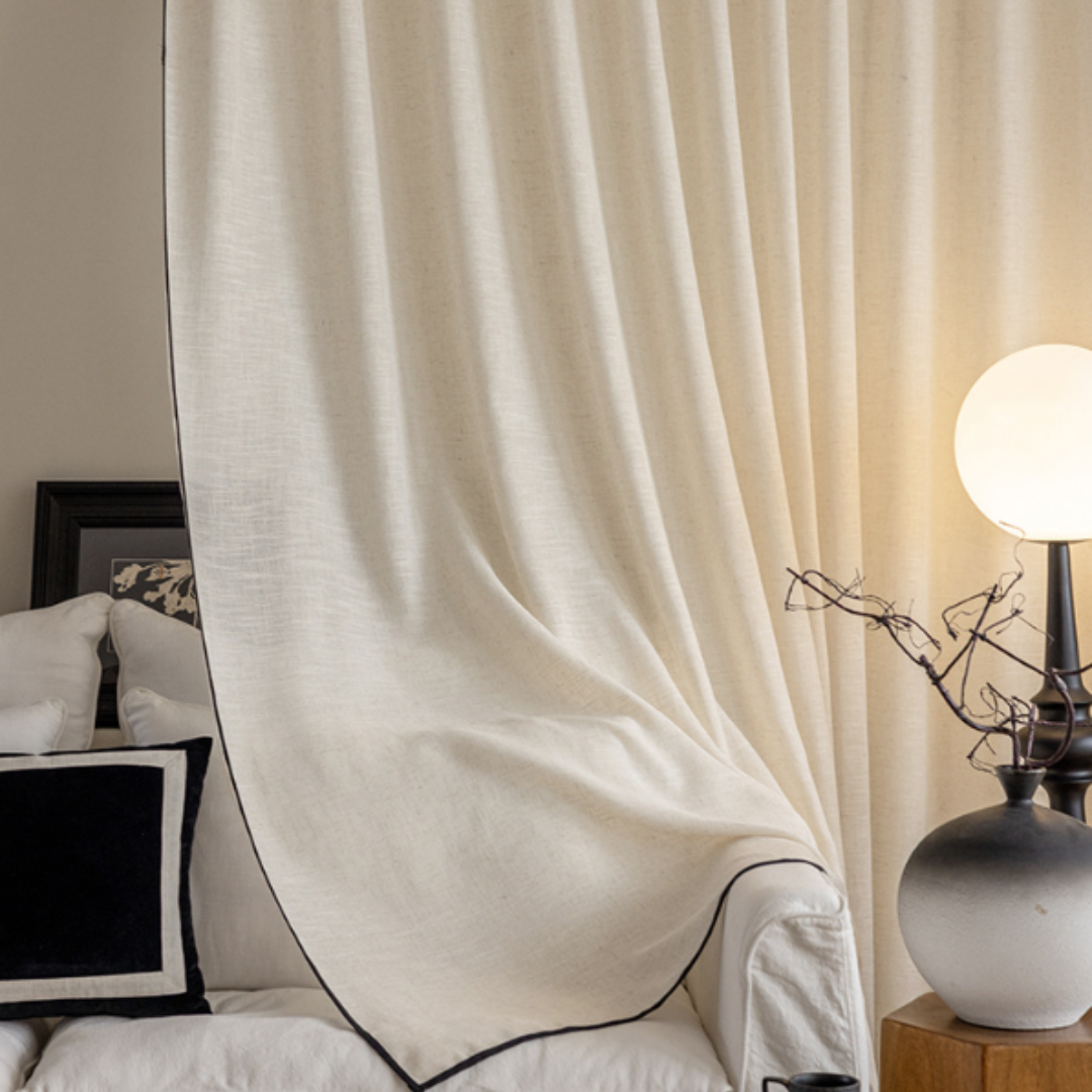 Beige linen curtain with black trim in a room setting with a lamp and vase.