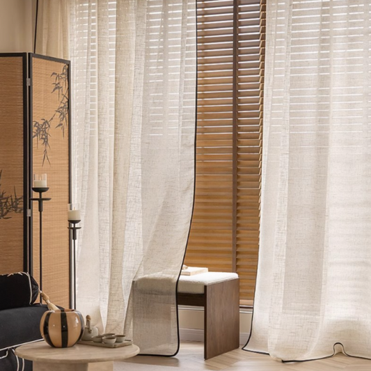Sheer linen curtains with black trim against wooden blinds in a room setting