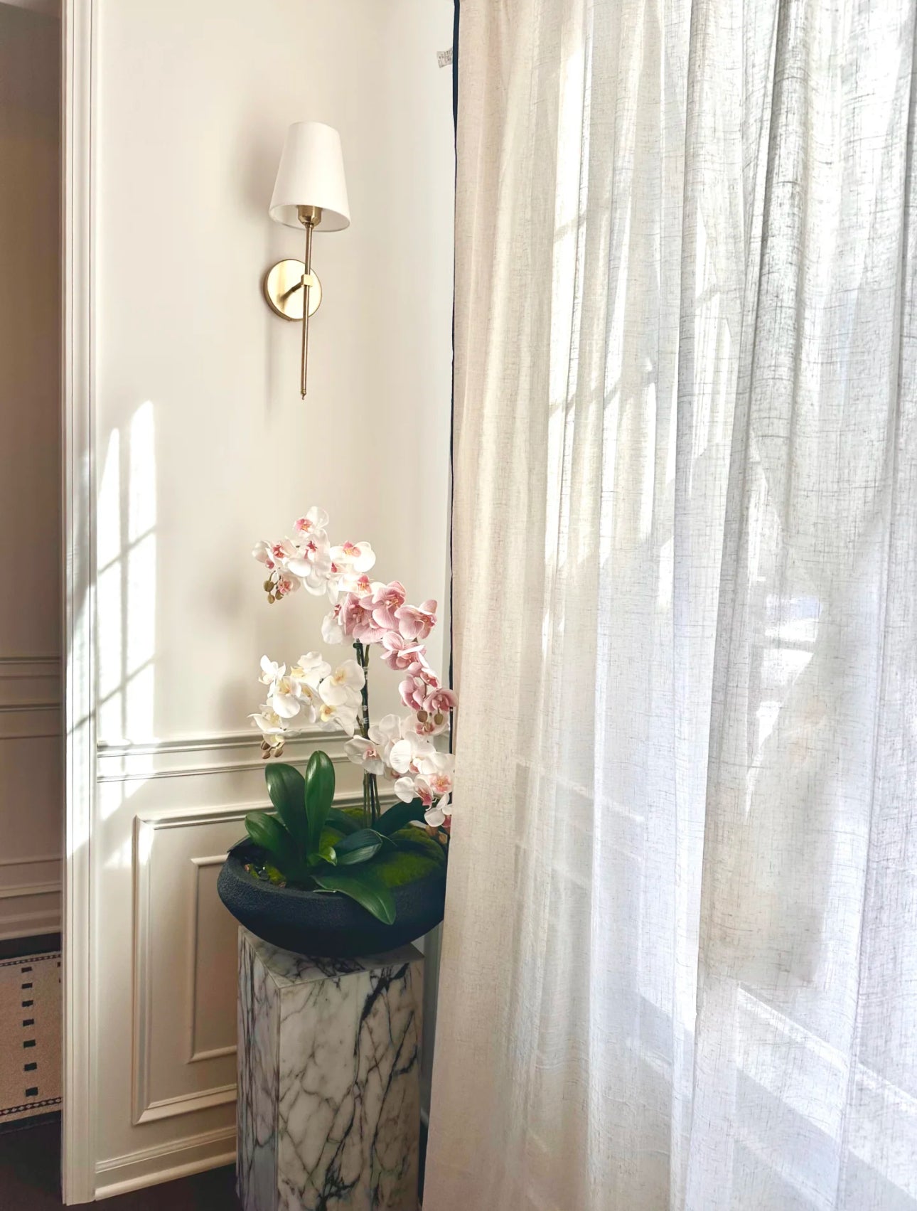 Potted orchid on a marble pedestal by a window with linen sheer curtains.