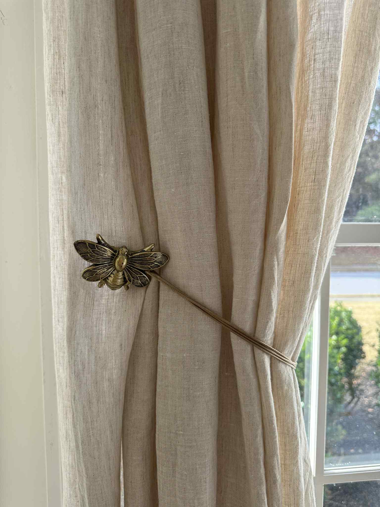 Beige curtain with a decorative bronze bee tieback in front of a window.