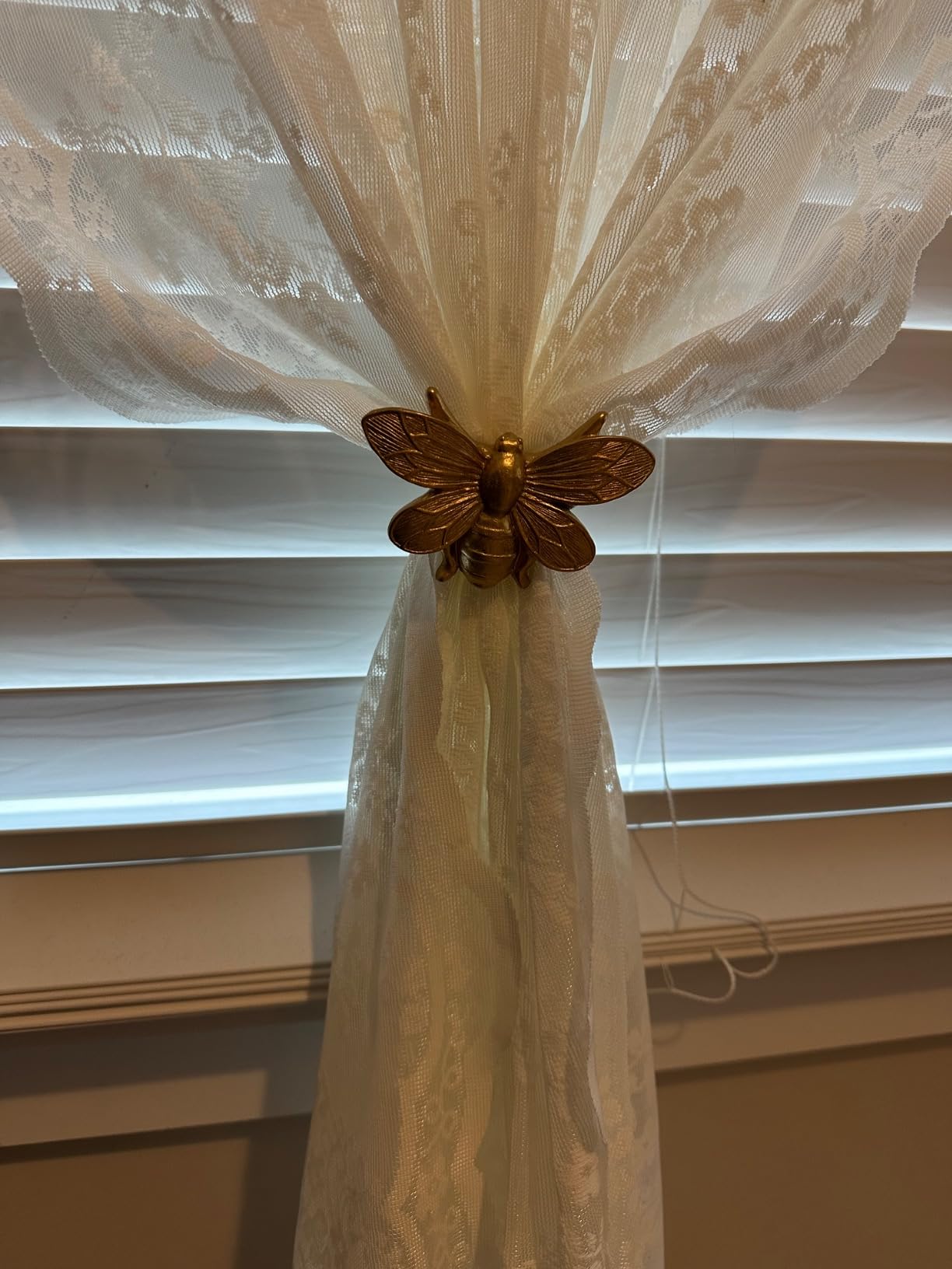 Decorative curtain tieback with a bumblebee design on a lace curtain in front of window blinds.
