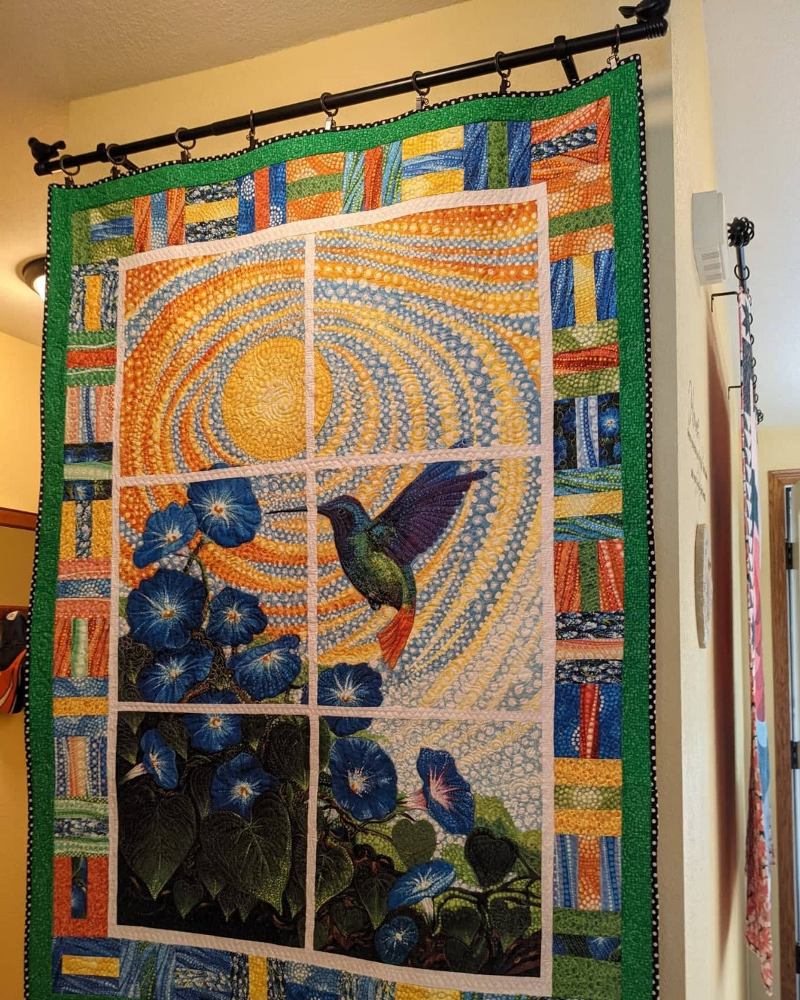 Colorful quilt with a bird and floral design hanging on a wall with a black curtain rod with bird ends