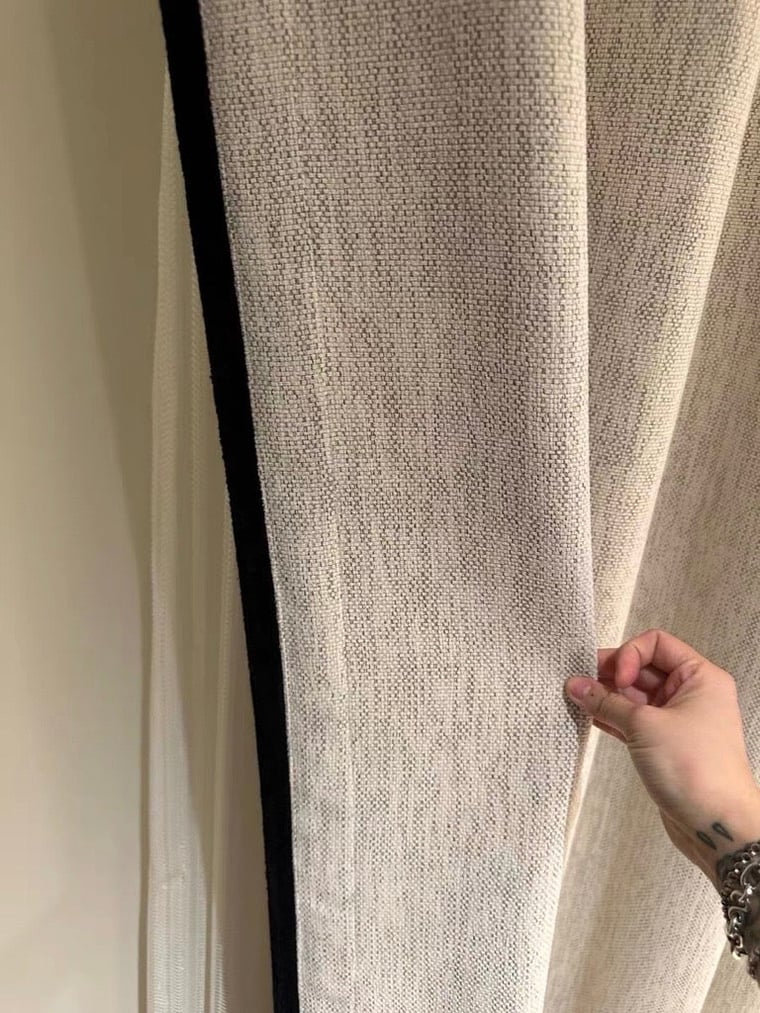 Close-up of a textured cream curtain with black trim with a hand holding it open.