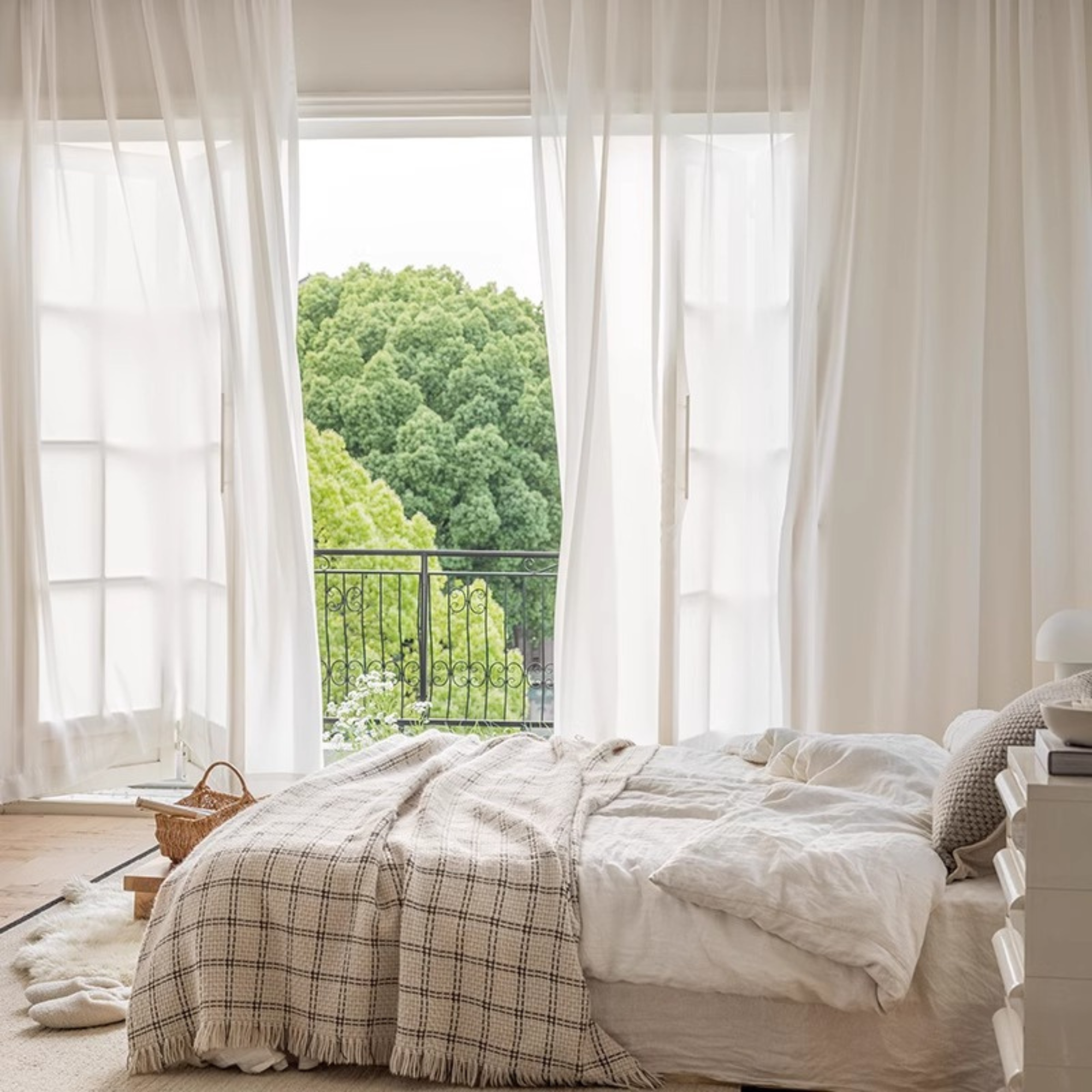 Neatly made bed with plaid blanket in a bright bedroom with white sheer curtains over sliding glass door.
