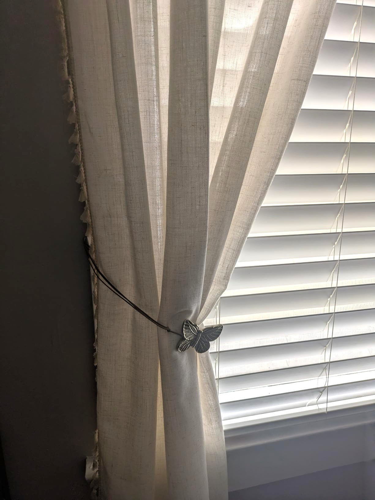 White curtain with a decorative butterfly tieback in front of a window with blinds.