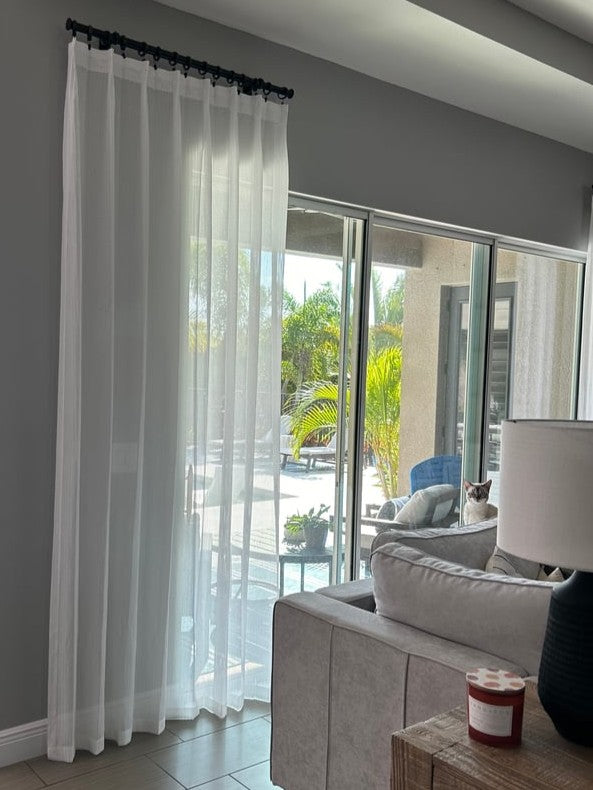Sheer white curtain in a living room with a view of a garden and a cat hiding behind couch.