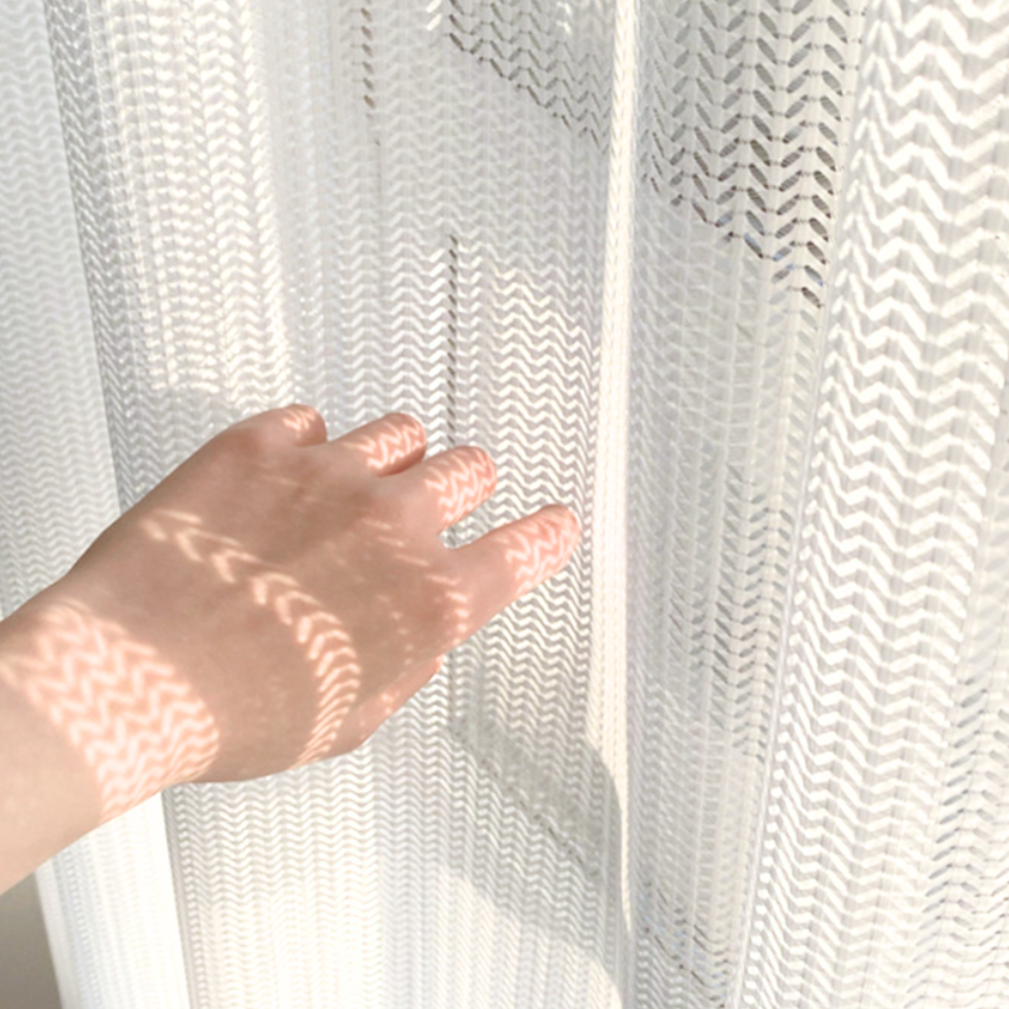Hand touching a textured white curtain with sunlight casting shadows, showing their delicate texture and cat-resistant weave.