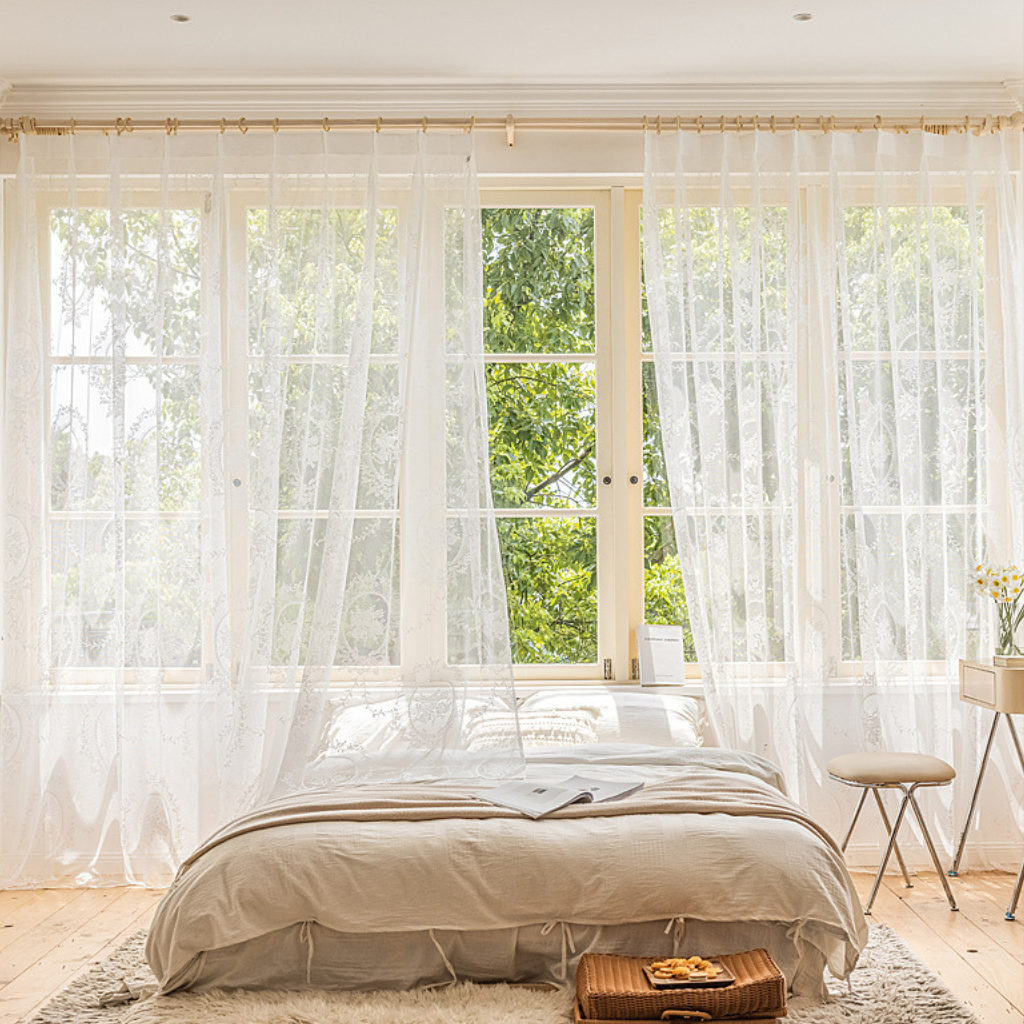 White sheer lace curtain panels hanging on a gold rod over a large multi-pane window in a bright, neutral-toned bedroom with a bed, small stool, and natural light.