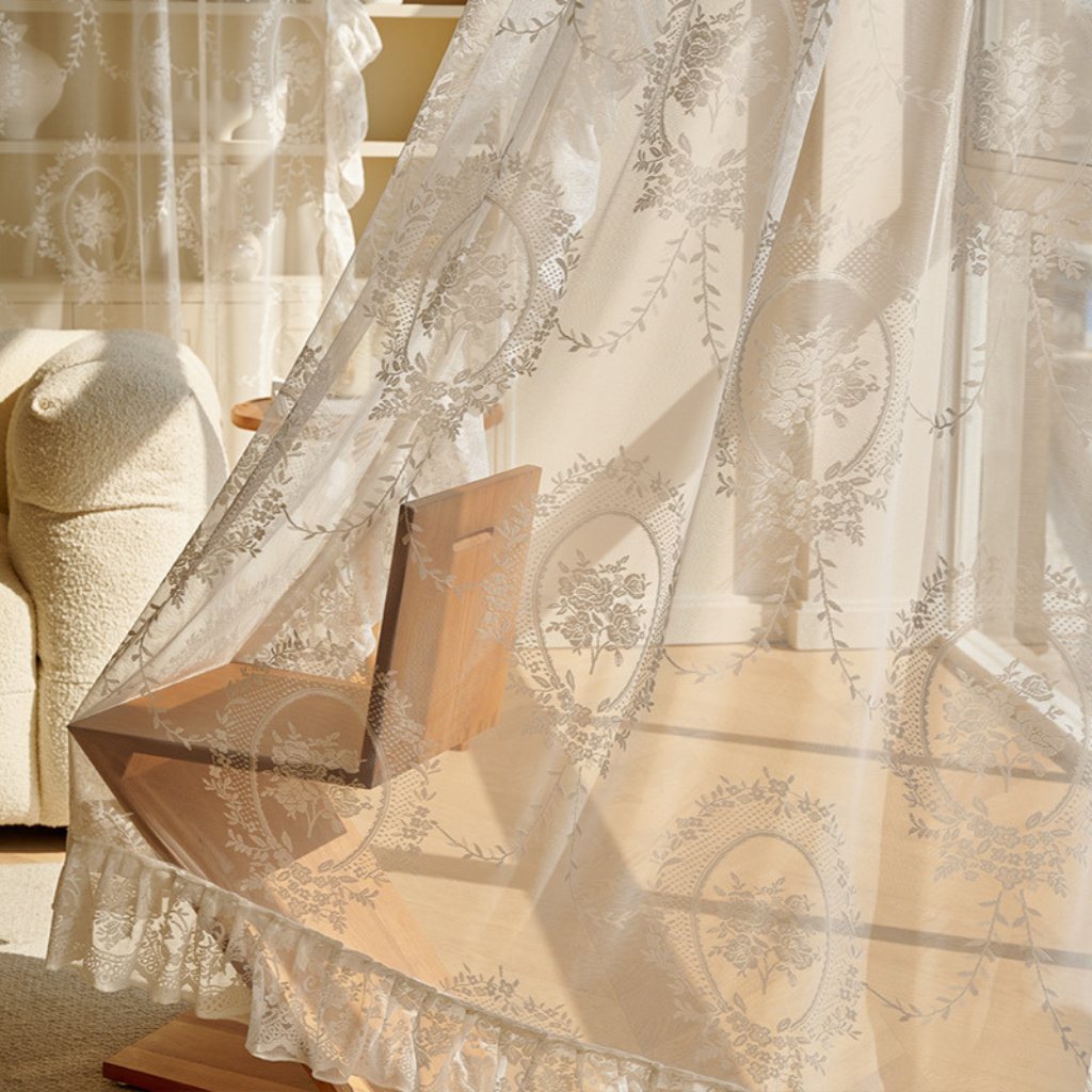 White sheer lace curtains with delicate floral embroidery and ruffled hems, shown hanging in a sunlit room with a wooden chair visible behind them.