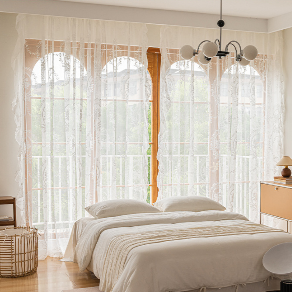 A bedroom scene with sheer white lace curtains hanging over tall arched windows, with a neatly made bed, bedside table, lamp, and chandelier visible in a softly lit interior.