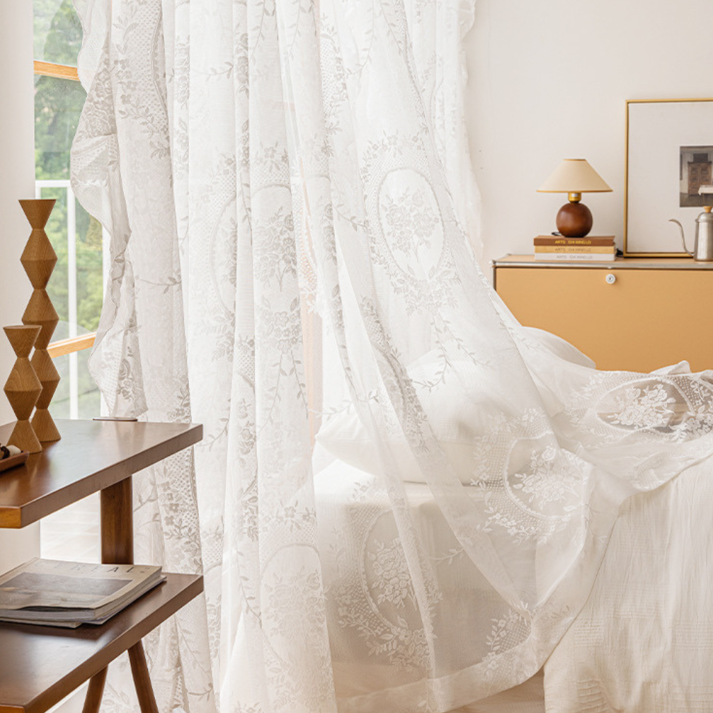 White sheer lace curtains with an intricate floral pattern hanging in a softly lit bedroom setting next to a wooden side table and bed.