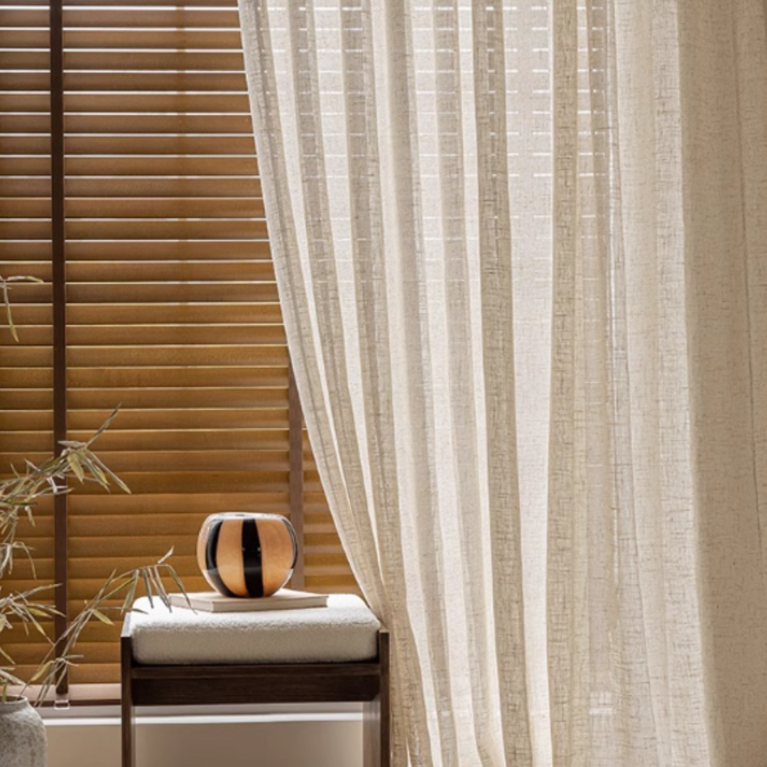 Sheer beige linen curtain with wooden blinds and decorative elements in a living room setting.