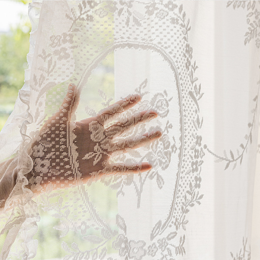 A close-up of a hand touching sheer white lace curtain fabric with floral embroidery and ruffled edges, with soft natural light filtering through.