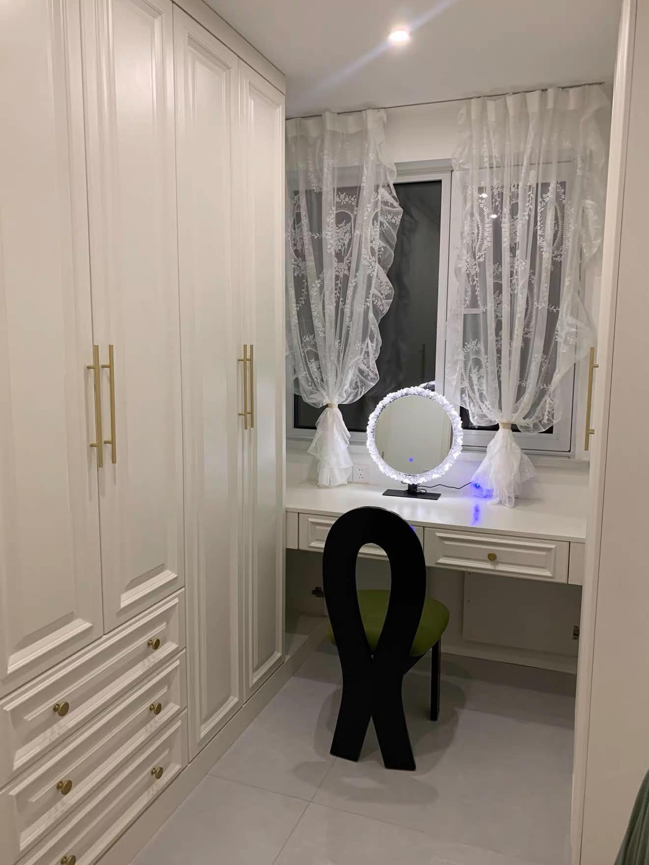 A white vanity desk setup in a room with tall white cabinets, sheer white lace curtains, a round mirror, and a black sculptural chair.