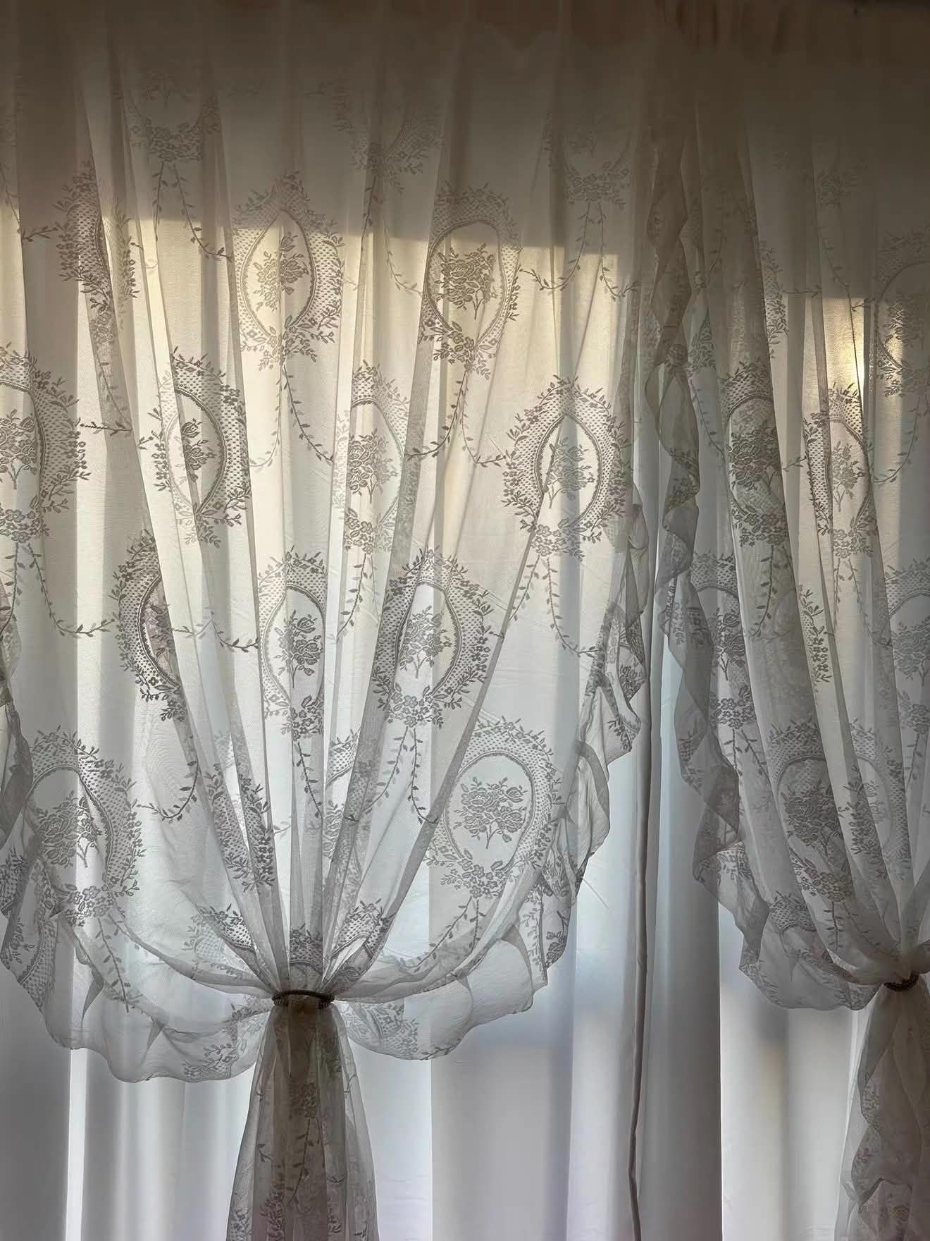 White sheer lace curtains with embroidered oval floral patterns, tied back to let light into a window.