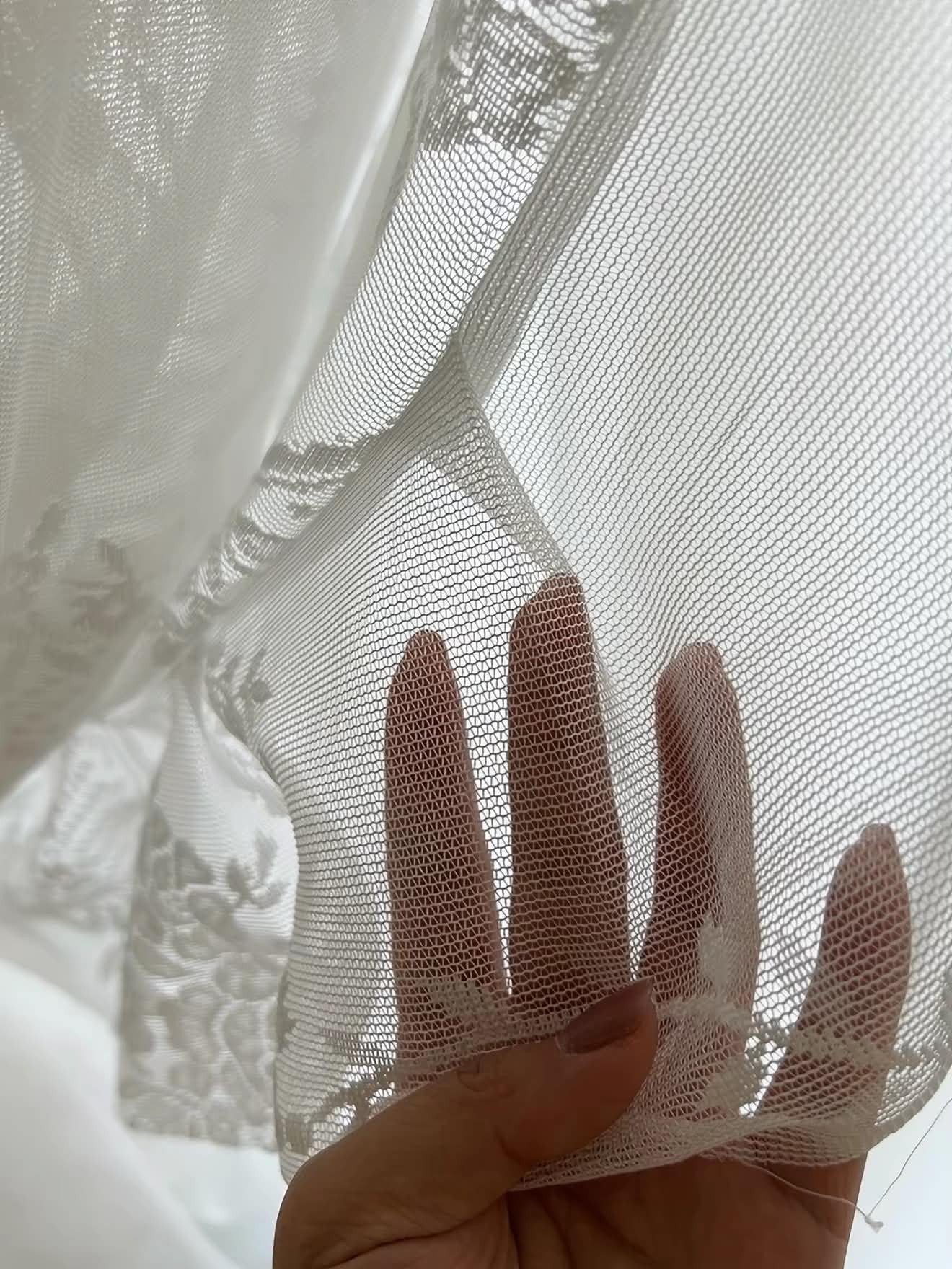 A close-up of a hand holding sheer white lace fabric with floral embroidery and mesh texture.