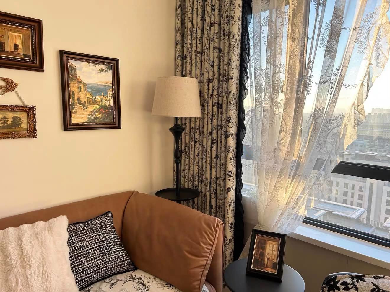 A cozy living room corner with a tan leather sofa, patterned pillows, a floor lamp, framed wall art, and a large window with sheer lace curtains and a city view.