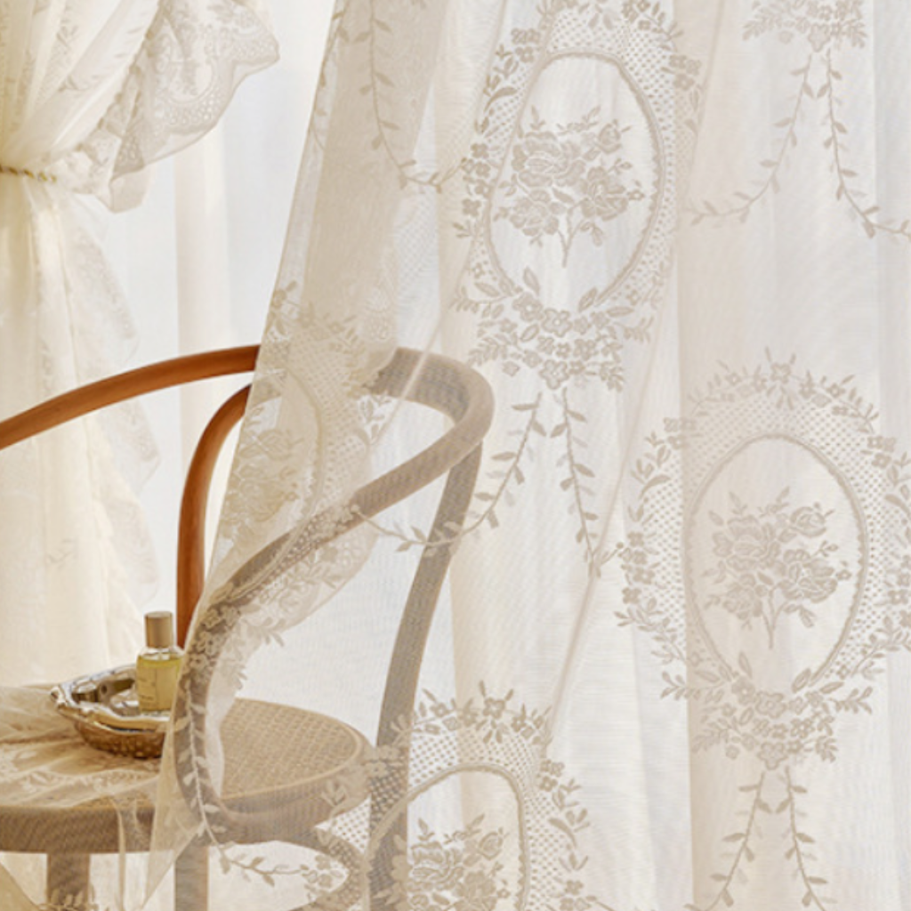 White sheer lace curtains with an elegant floral and oval cameo pattern, draped over a wooden chair in a softly lit room.