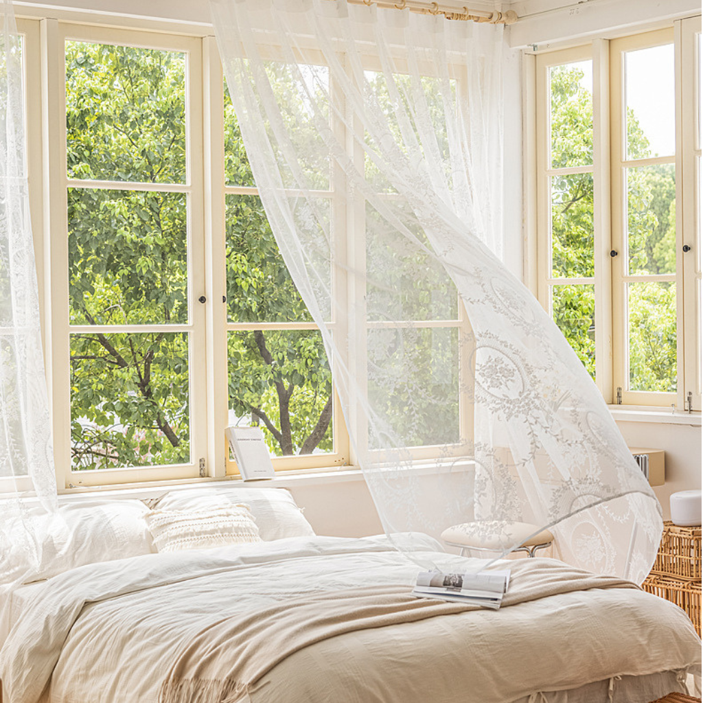 A beautifully styled bedroom with a bed dressed in soft beige linens, sheer white lace curtains billowing in the wind, and large windows looking out onto lush green trees.
