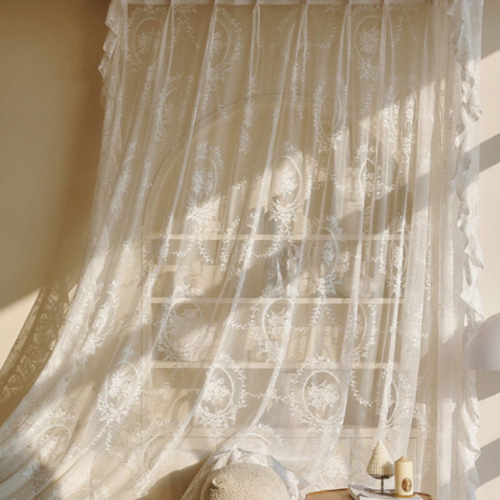 White embroidered sheer curtain panels with floral and oval lace patterns, hanging in front of a window and draping over a bed, with warm sunlight casting shadows.