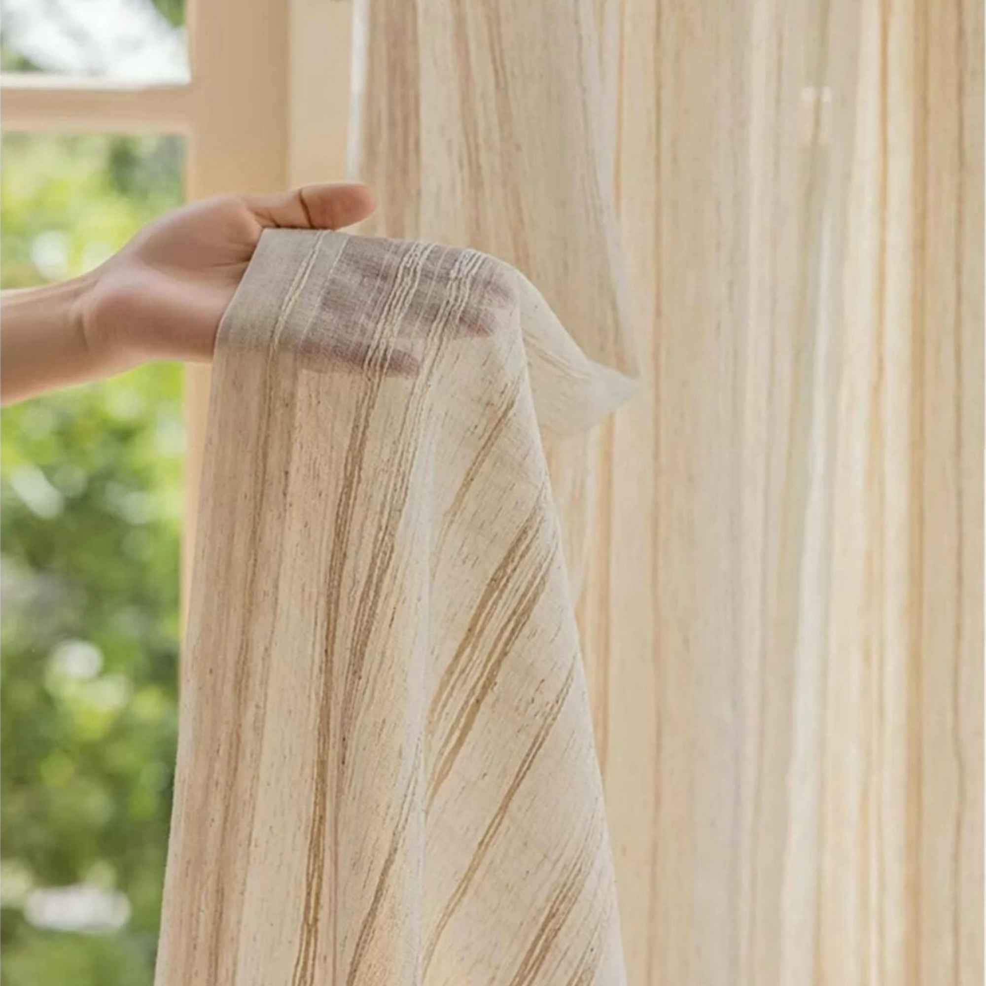 Hand holding a piece of beige linen curtain fabric with subtle stripe patterns against a blurred natural background