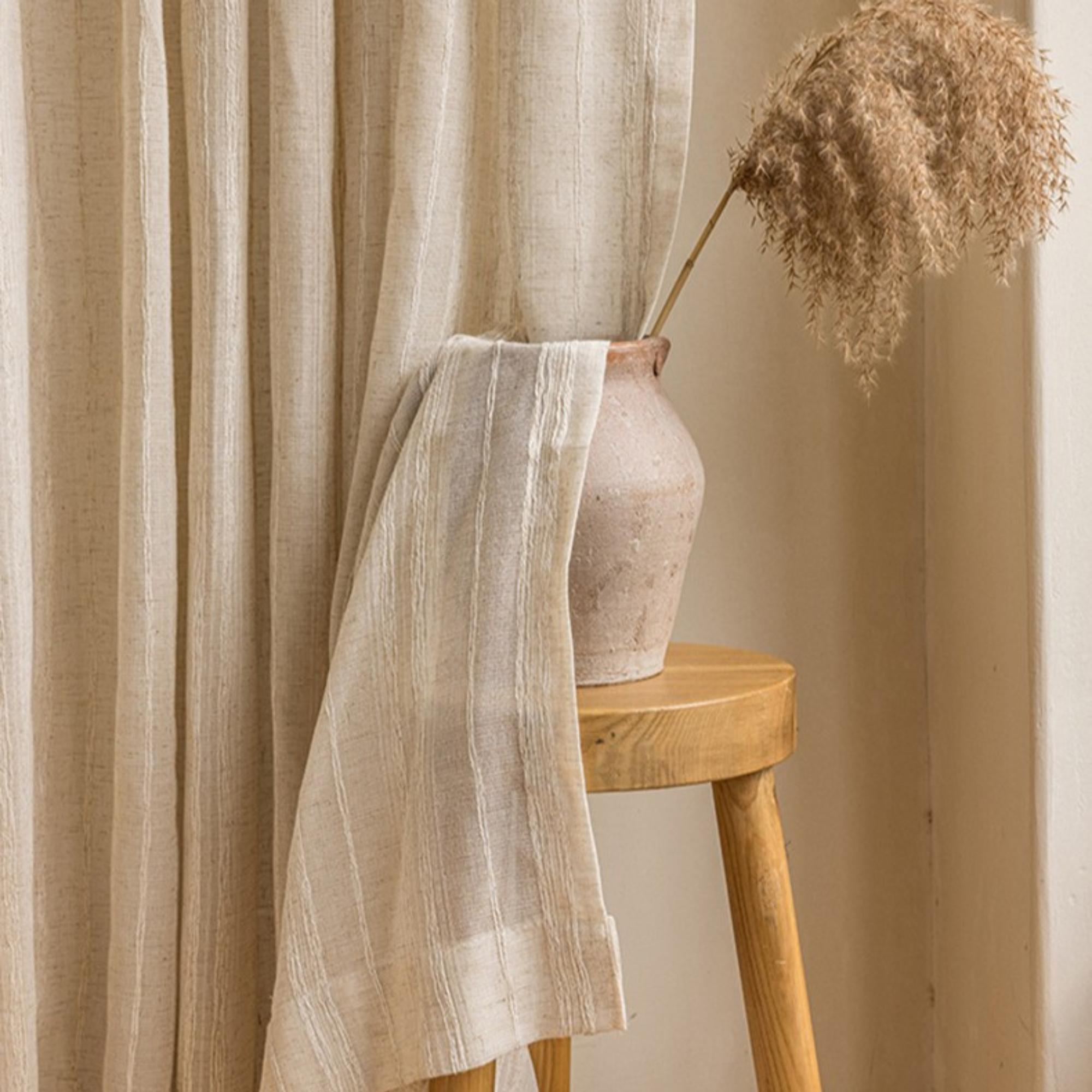 Beige sheer linen curtain with a vase and dried plant on a wooden stool against a beige wall